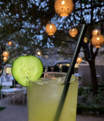 a margarita with a cucumber garnish is in front of large outdoor lights hanging from trees on a patio with  outdoor umbrellas over the tables