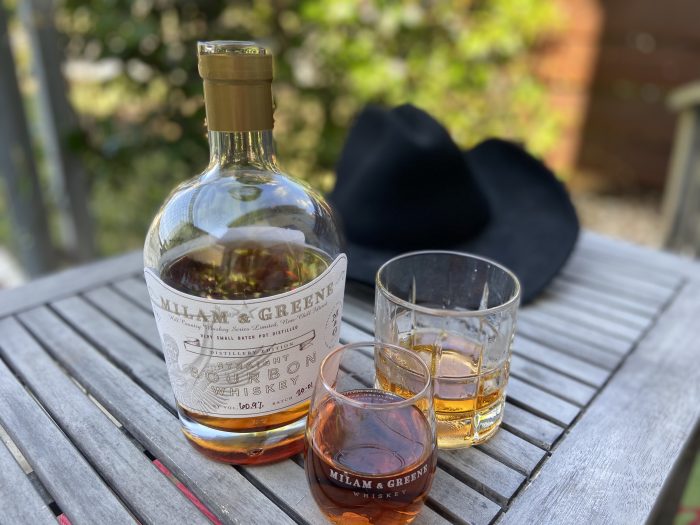 a bottle of whiskey is on a table with a black cowboy hat behind it and two poured glasses of whiskey in front of the bottle