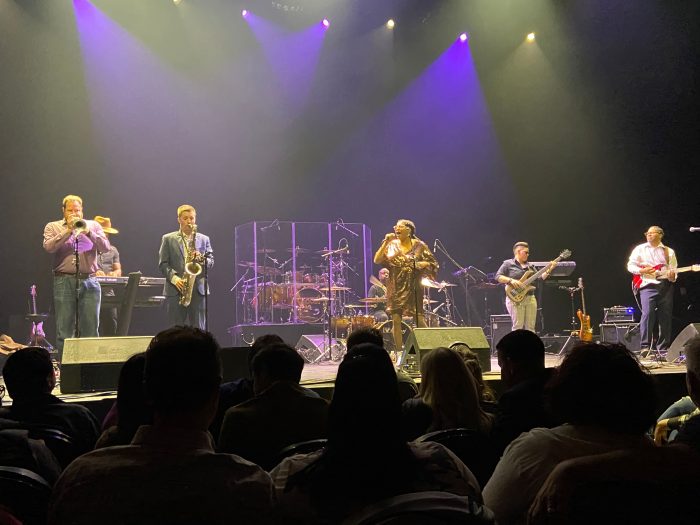a 7-piece band is on a stage lit up with purple and blue lights, and a young African-American woman in a shiny gold dress is singing
